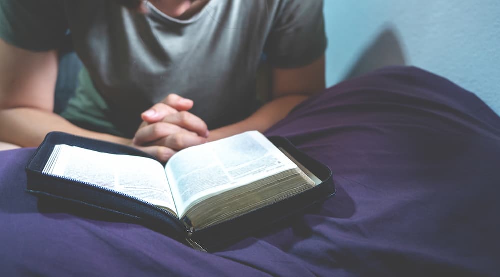 Man kneeling in front of bed praying with Bible